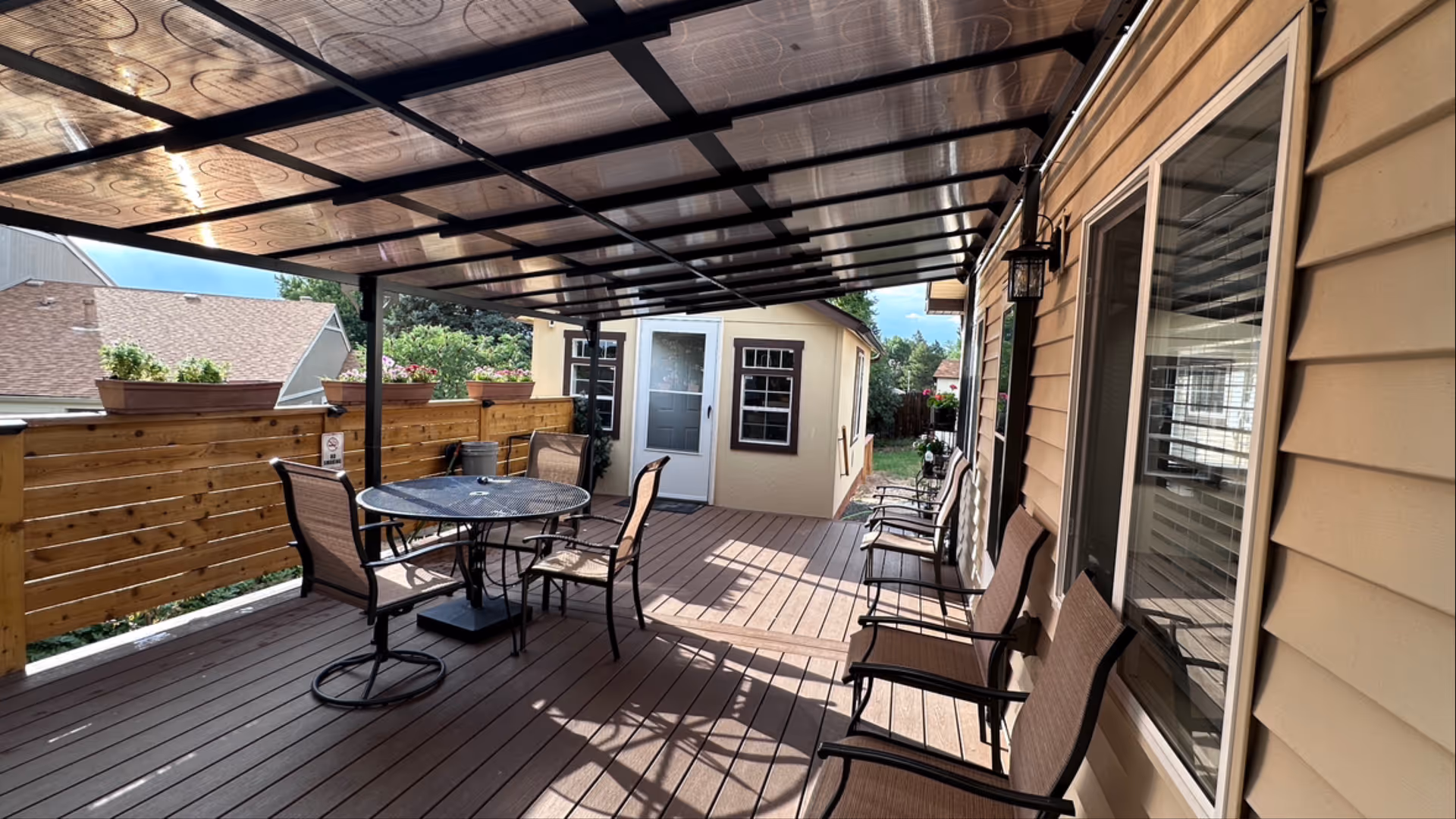 Covered outdoor patio area with a transparent roof, featuring a round table with four chairs and additional chairs lined up against the house wall. The patio has wooden flooring and a wooden fence with flower pots on top. A small building with a door and windows is visible at the end of the patio.
