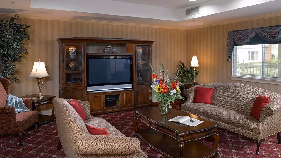 A cozy living room with two patterned sofas and a matching armchair, all adorned with red throw pillows. A wooden coffee table with a vase of colorful flowers and an open book sits in the center. Against the wall is a large wooden entertainment center with a flat-screen TV. The room has warm beige striped wallpaper, a red patterned carpet, two table lamps, a large window with floral valance, and some green plants.