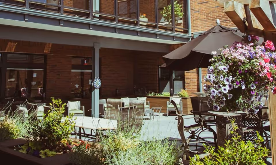 Sunlit courtyard patio with tables, chairs, umbrellas, and flowering planters in front of a brick building.