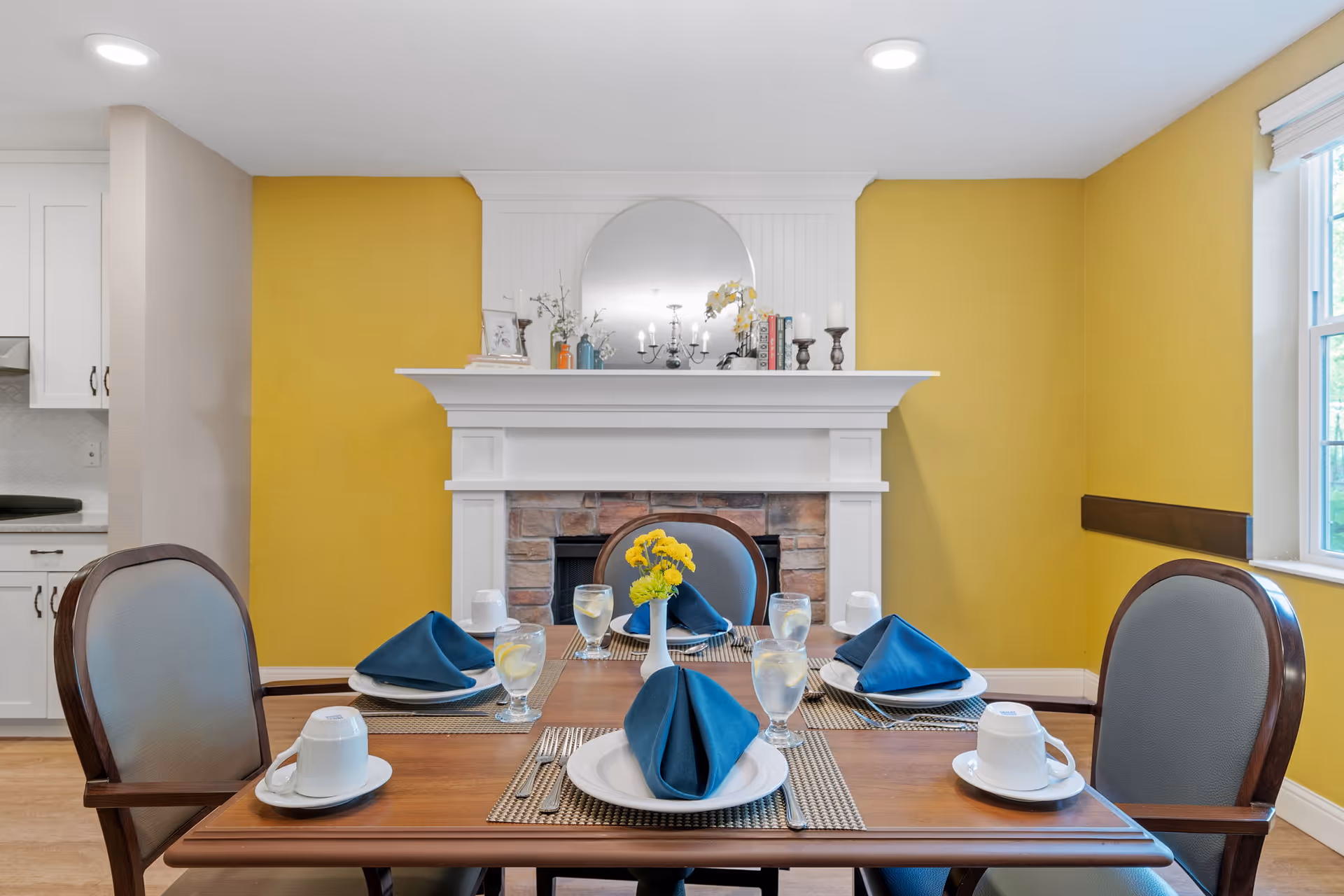 A dining room with a wooden table set for four people with white plates, blue folded napkins, glasses of water with lemon, and white cups. The room features a white fireplace with a round mirror above it, decorative items on the mantel, and yellow walls. There are four upholstered chairs around the table and a window on the right side letting in natural light.