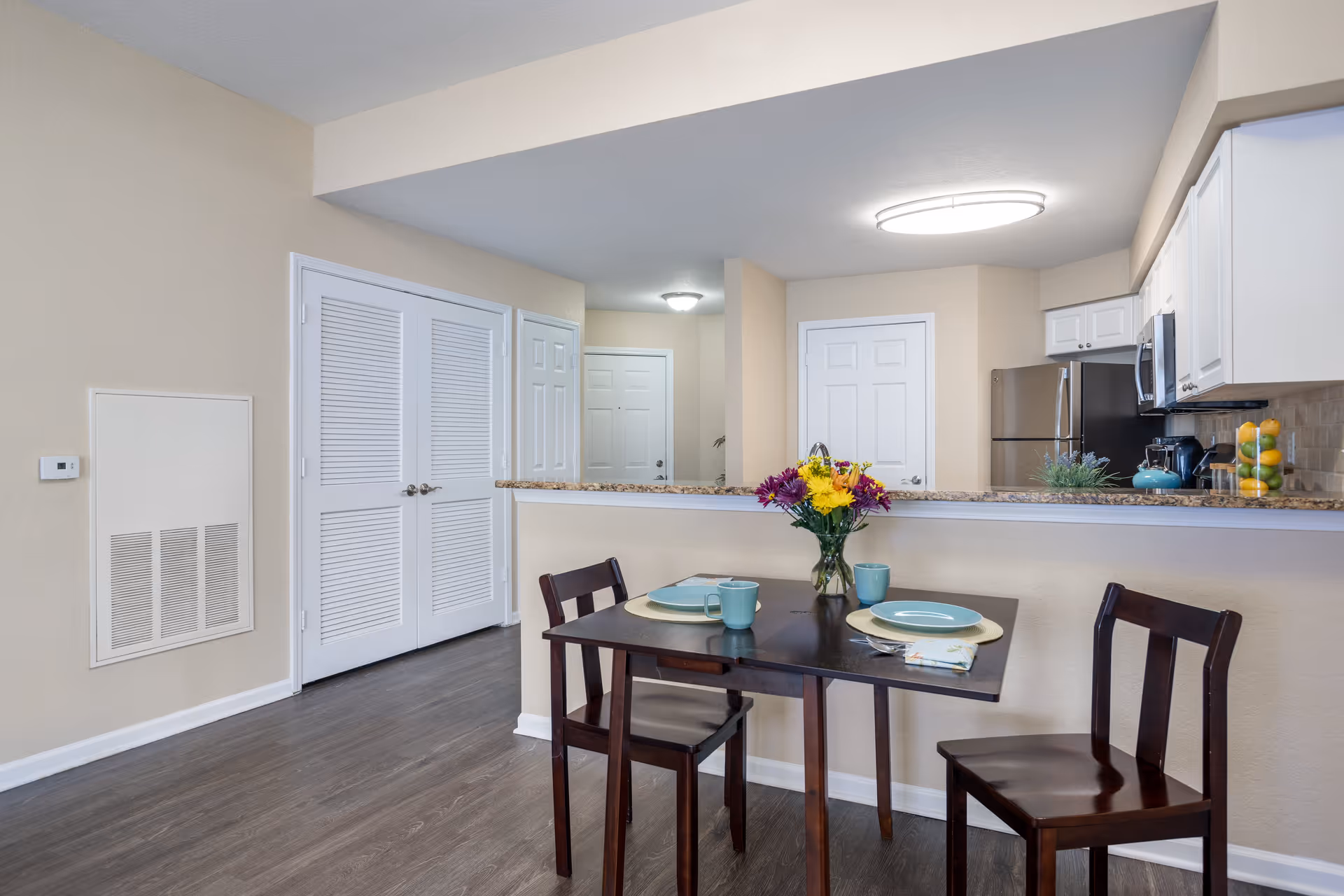 A small dining area with a dark wooden table set for two with plates, mugs, and a vase of colorful flowers. The dining area is adjacent to a kitchen with white cabinets, a stainless steel refrigerator, and a granite countertop. The walls are beige, and the floor is dark wood. There are two closed white doors and a thermostat on the wall.