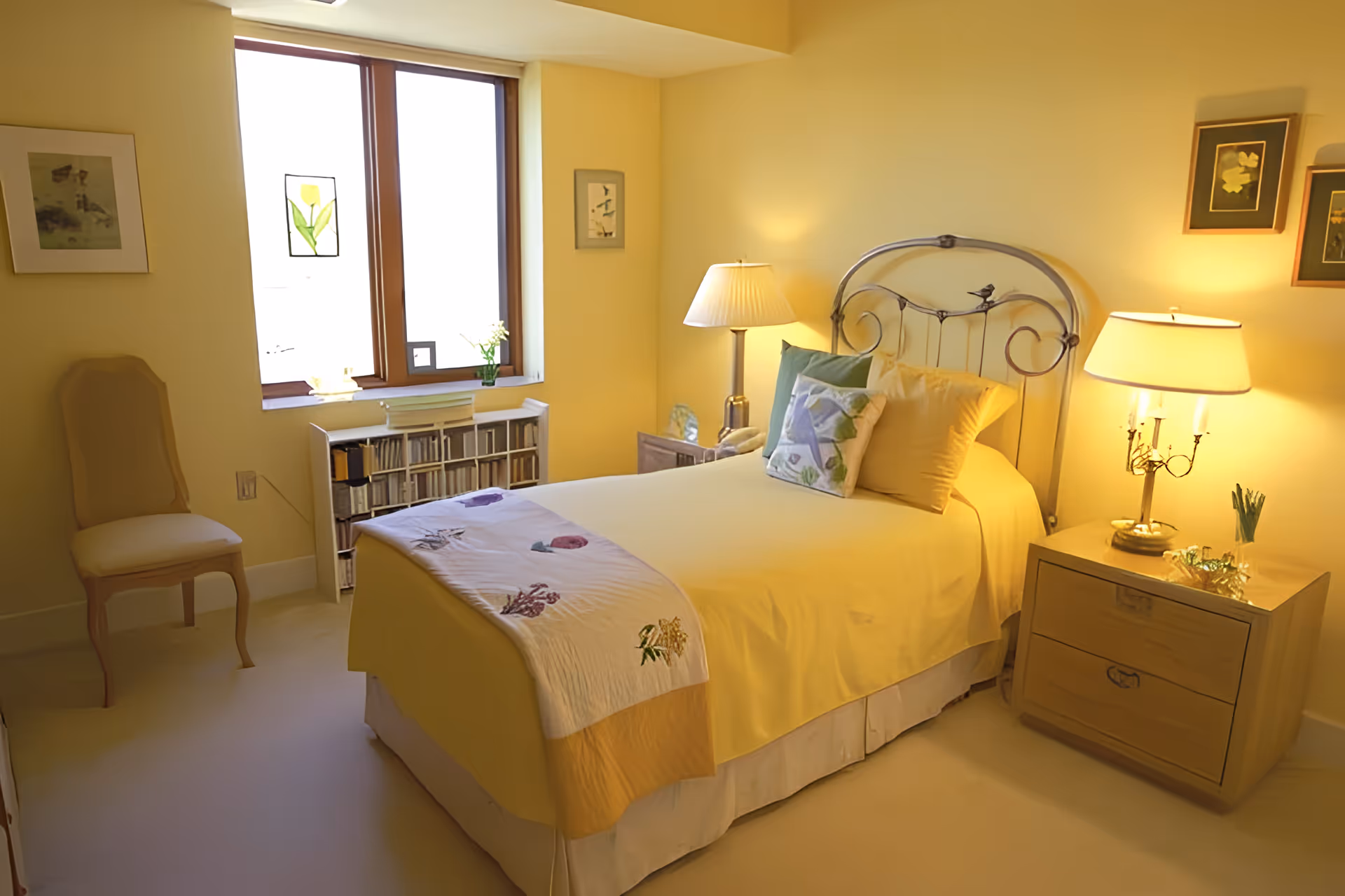 Bright yellow-themed bedroom with a single bed, metal headboard, bedside tables with lamps, a chair and a window.