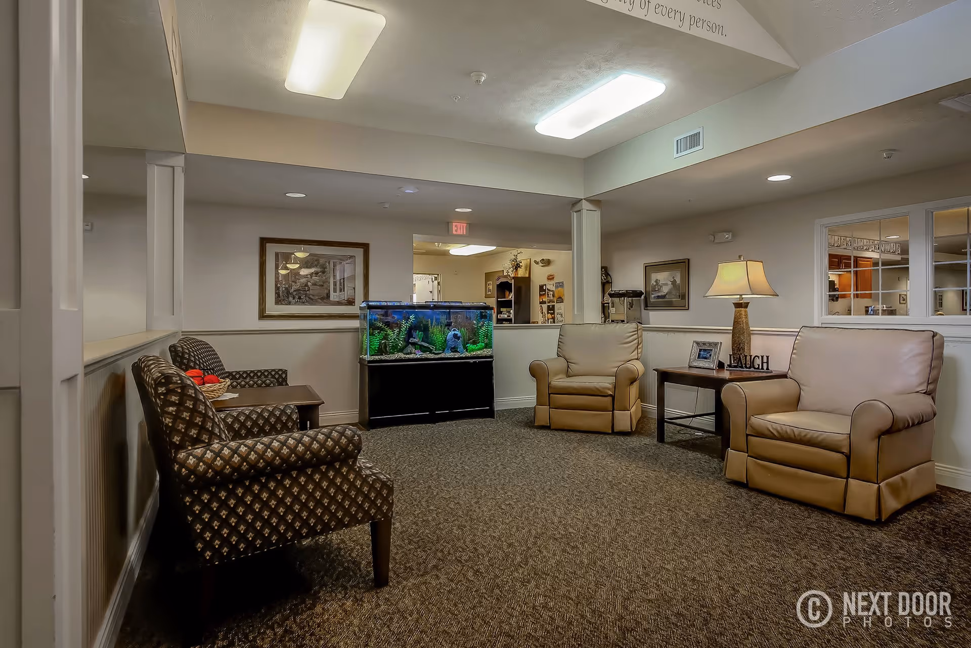 A cozy living room area in Grandhaven Living Center featuring two patterned armchairs on the left, two beige leather armchairs on the right, a wooden side table with a lamp and decorative items, and a large fish tank against the back wall. The room has carpeted flooring, neutral-colored walls, framed artwork, and ceiling lights.