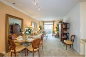 A well-lit living and dining area in a senior living facility. The dining area features a round glass table set with plates, glasses, and a floral centerpiece, surrounded by four wooden chairs. The living area has a large window with curtains, a sofa, a coffee table, and a tall shelving unit with decorative items. The walls are painted in neutral tones, and a large mirror hangs above a wooden sideboard.