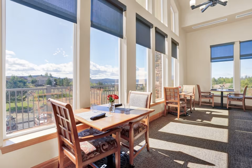 Bright dining area with wooden tables and cushioned chairs next to large windows with blue roller shades, overlooking a scenic outdoor view with trees and hills under a partly cloudy sky.