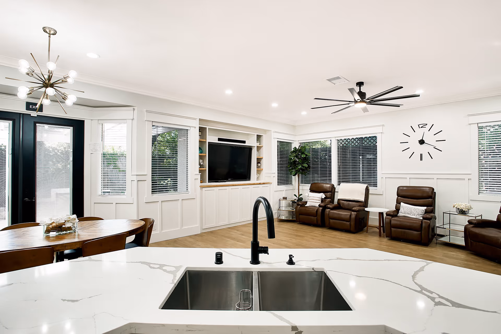 Modern senior living common area with a white marble countertop and sink in the foreground, a wooden dining table with chairs to the left, and a living room area with brown leather recliners, a large wall clock, a ceiling fan, and a flat-screen TV mounted on a white built-in cabinet. Large windows with blinds and glass doors provide natural light.