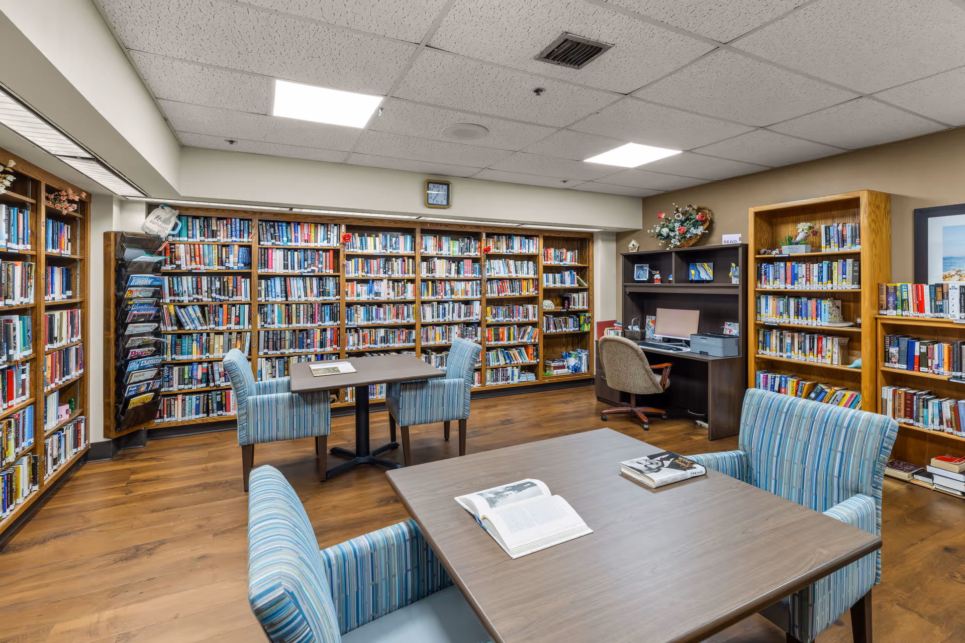 A well-lit senior living facility library room with wooden bookshelves filled with books lining the walls. There are two tables with blue striped upholstered chairs around them, and an open book and a closed book on the nearest table. A computer desk with a chair and a printer is positioned against the wall on the right side. The room has a wooden floor and a drop ceiling with fluorescent lights.
