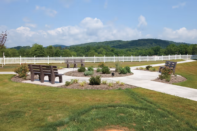 Outdoor garden area with paved walkways forming a circular pattern around landscaped flower beds and shrubs. Several wooden benches are placed along the walkways. A white picket fence borders the area, with green trees and hills visible in the background under a partly cloudy sky.
