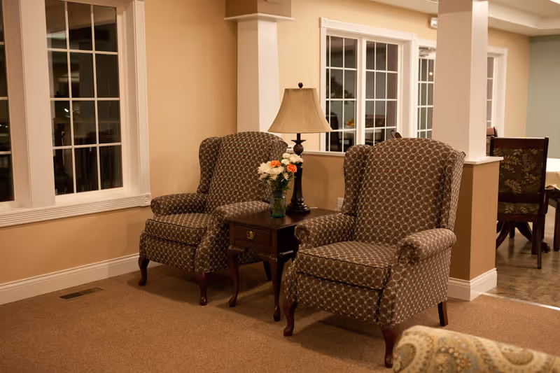 A cozy seating area with two patterned armchairs separated by a small wooden side table holding a lamp and a vase of flowers. The space has beige walls, carpeted floor, and large windows with white trim. In the background, a dining area with chairs and a table is partially visible.