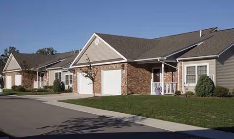 Row of single-story residential buildings with garages, brick and siding exteriors, small front porches, and well-maintained lawns under a clear blue sky.