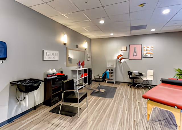 Interior view of a room with light gray walls and wood-patterned flooring. The room contains a black chair in front of a mirror with wall-mounted lights, a black sink on the left, shelves with towels and supplies, a red cushioned examination or massage table on the right, and a small desk with chairs and artwork on the far wall.