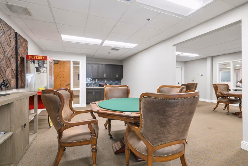 A well-lit room with a round green felt card table surrounded by four brown leather and wood chairs. In the background, there is a popcorn machine and a kitchenette with dark cabinets and a tiled backsplash. The room has beige carpet and white walls with a drop ceiling and fluorescent lighting.