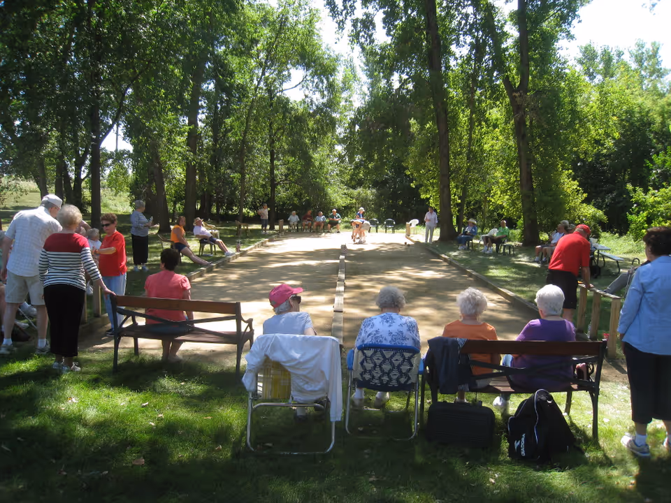 A group of seniors seated around a tree-lined outdoor bocce court while players compete.