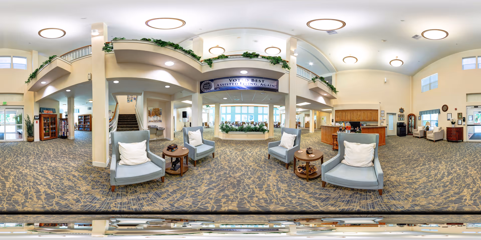 A spacious and well-lit senior living facility lobby with comfortable seating including armchairs and small tables. The area features a carpeted floor with a patterned design, a reception desk on the right side, and a staircase leading to an upper level. Green plants decorate the balcony railing above, and large windows allow natural light to fill the space. A banner above the reception area reads 'Voted Best Assisted Living Again.'