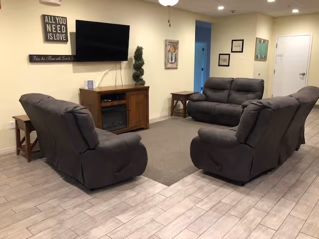 A cozy living room area with four gray recliner sofas arranged around a small wooden entertainment center with a flat-screen TV mounted above it. The walls are light yellow with framed artwork and inspirational signs, including one that says 'ALL YOU NEED IS LOVE'. The floor is covered with light wood-like tiles and a gray area rug in the seating area.