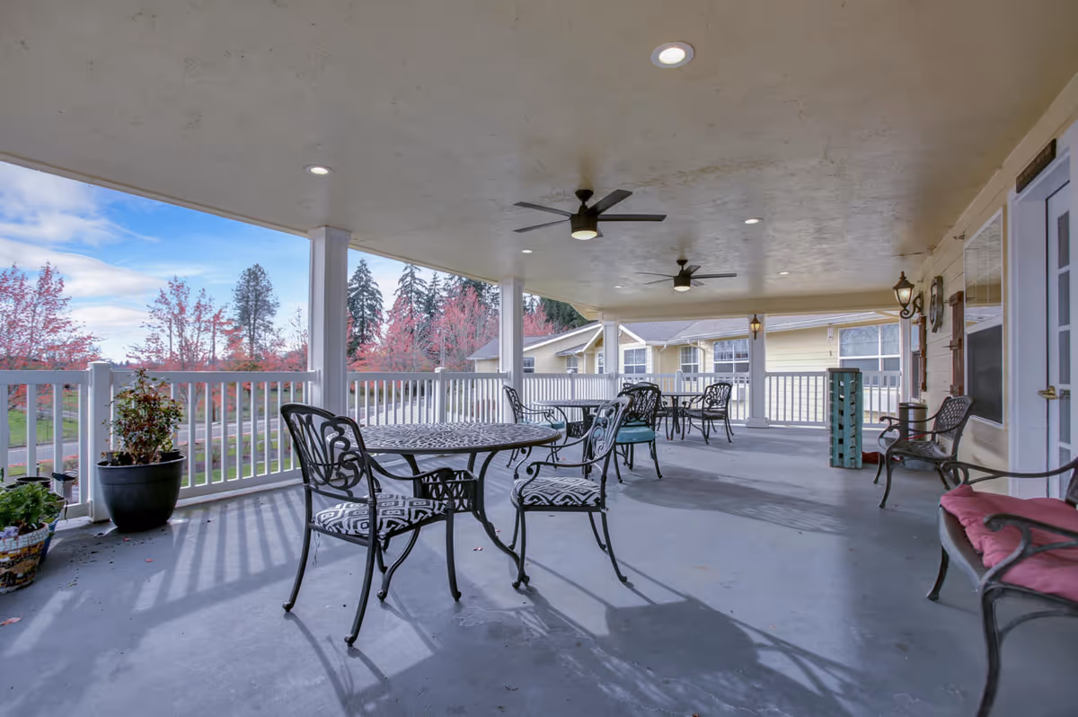 Covered outdoor patio with metal tables and chairs, ceiling fans, potted plants, and a white railing overlooking landscaped grounds.