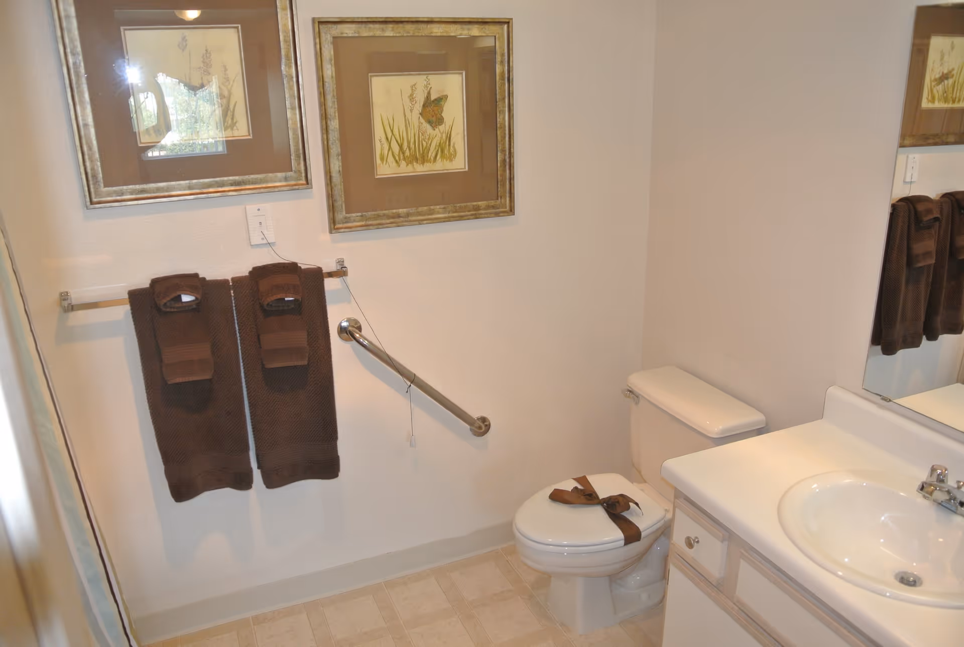 A small bathroom with a white toilet and sink. Two brown towels hang on a towel rack on the wall above a metal grab bar. Two framed pictures of nature scenes are hung above the towels. The toilet seat is secured with a brown strap. The floor has light-colored tiles.