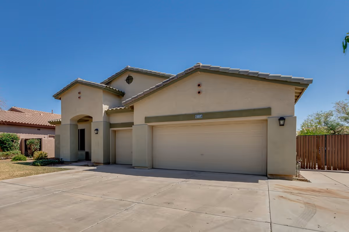 Exterior view of a single-story residential building with a beige facade, tiled roof, and a large garage door. The driveway is concrete, and there is a small patch of grass and shrubs on the left side. The sky is clear and blue.