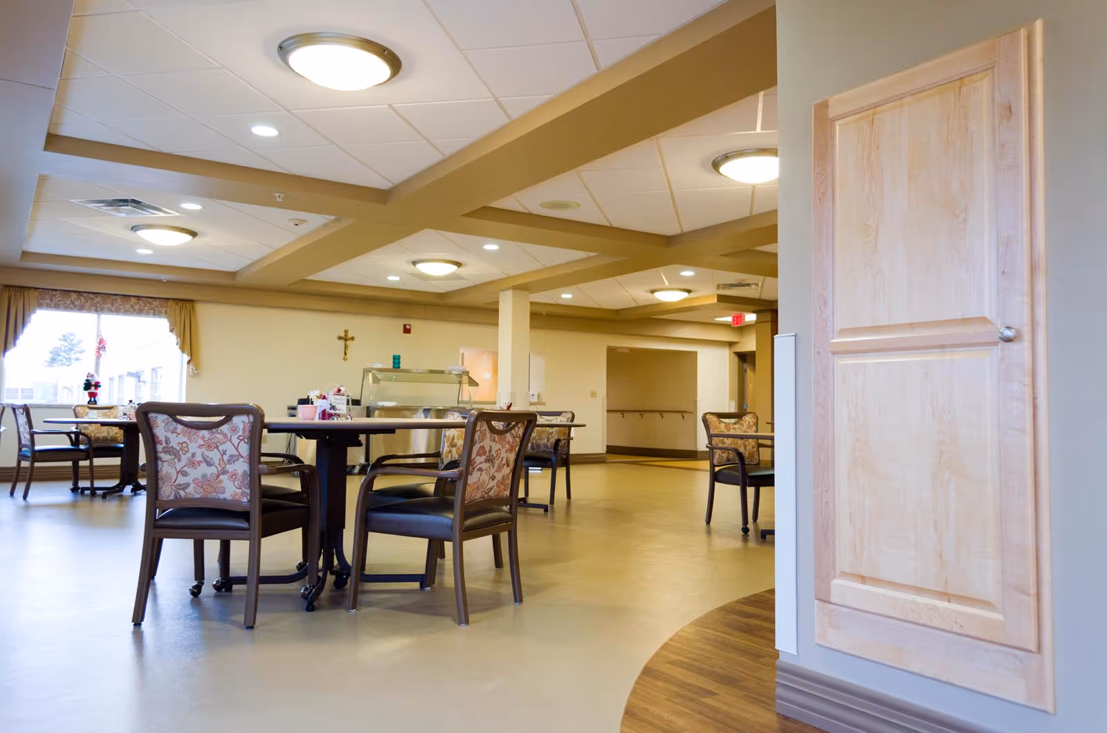 Bright communal dining area with round tables and floral-upholstered chairs and a serving counter in a senior living facility.