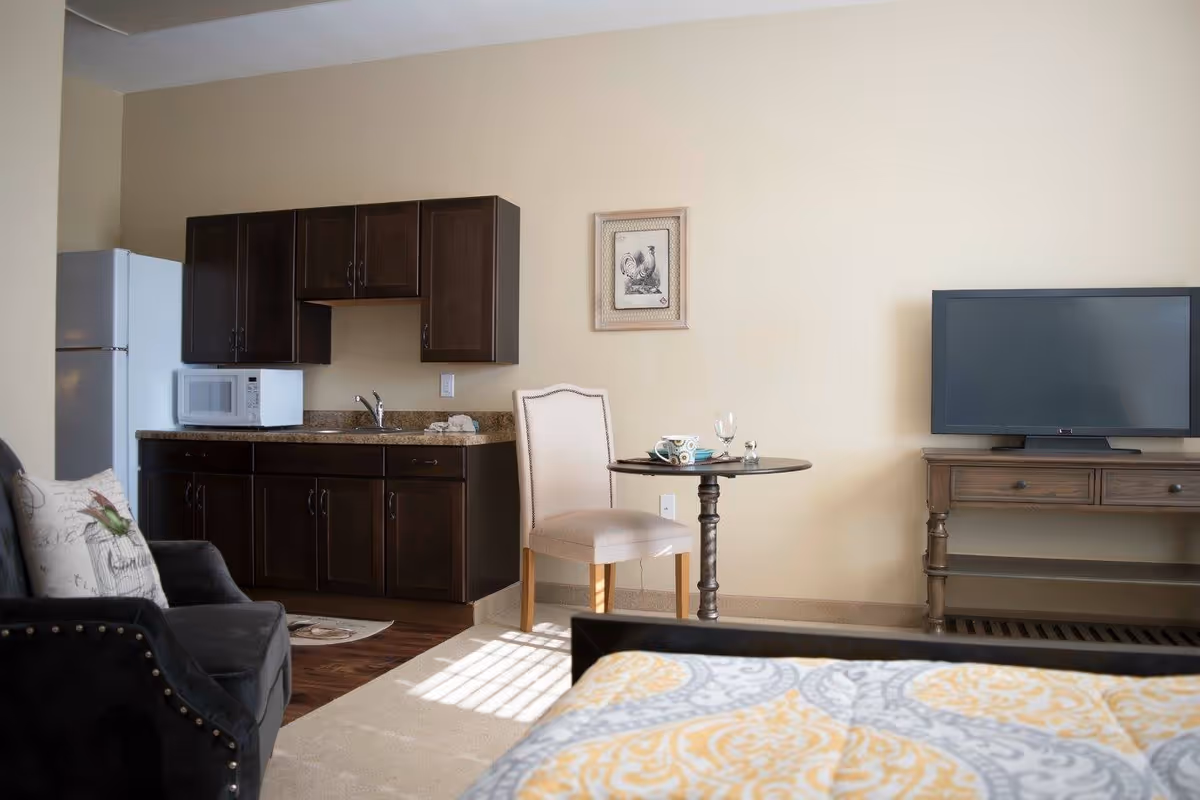 A cozy senior living studio apartment featuring a kitchenette with dark wood cabinets, a white refrigerator, and a microwave. There is a small round table with a chair, a flat-screen TV on a wooden stand, a black armchair with a decorative pillow, and part of a bed with a patterned yellow and gray bedspread visible in the foreground.