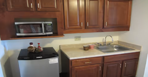 A small kitchen area with wooden cabinets, a countertop with a sink, a microwave mounted above a mini refrigerator, and some decorative items on the counter and refrigerator.