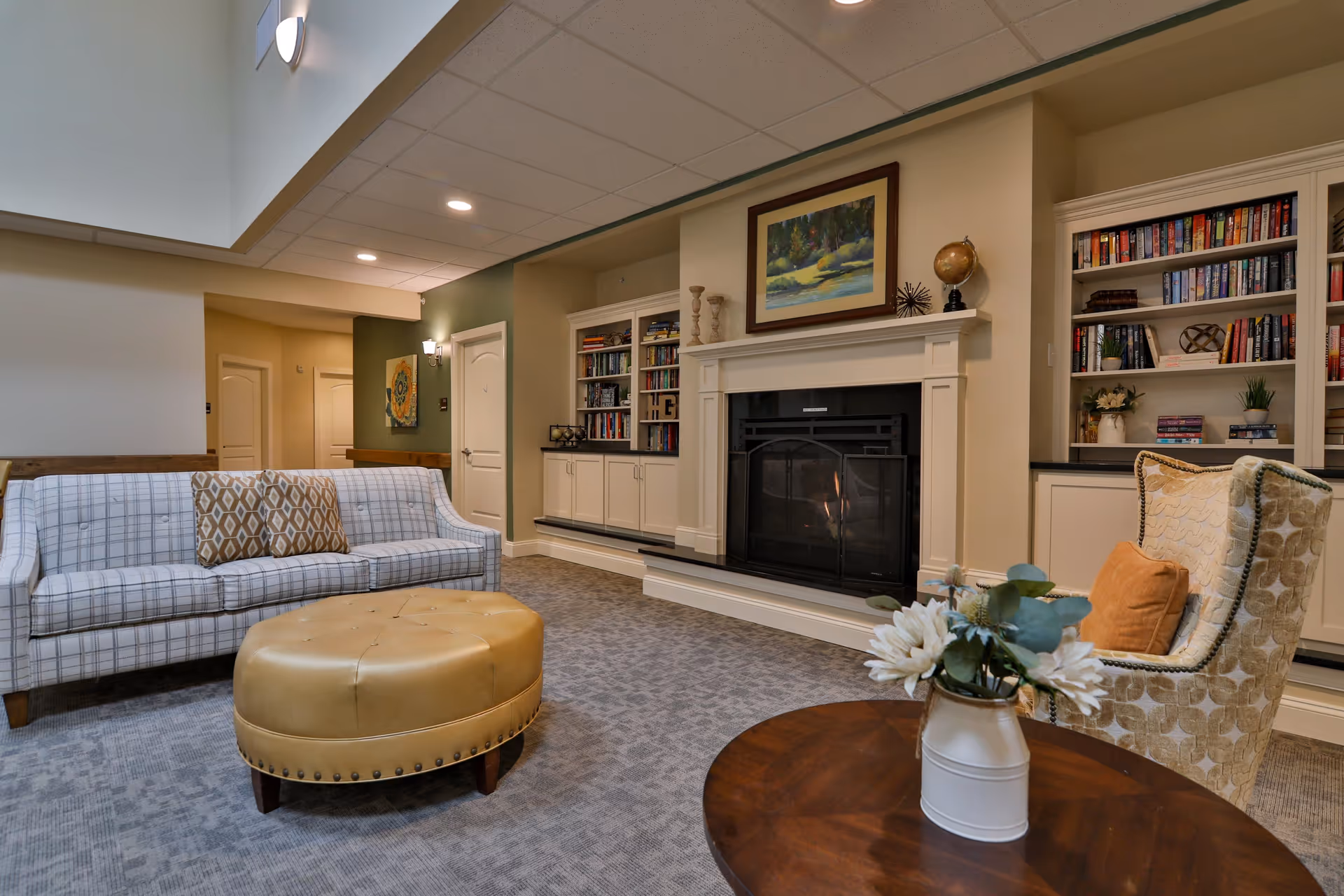 A cozy senior living common area with a plaid sofa, a round tan ottoman, a patterned armchair with an orange pillow, a wooden coffee table with a white vase of flowers, built-in bookshelves filled with books, and a fireplace with a painting above it.