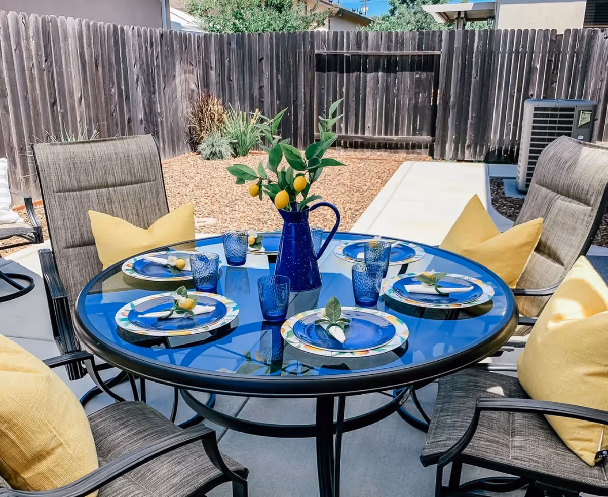 Outdoor patio area with a round glass table set for six, featuring blue plates, blue glasses, and yellow napkins with decorative flowers. The table has a blue pitcher with green leaves and yellow lemons as a centerpiece. Surrounding the table are cushioned chairs with yellow pillows. The background shows a wooden fence, some plants, and an air conditioning unit.