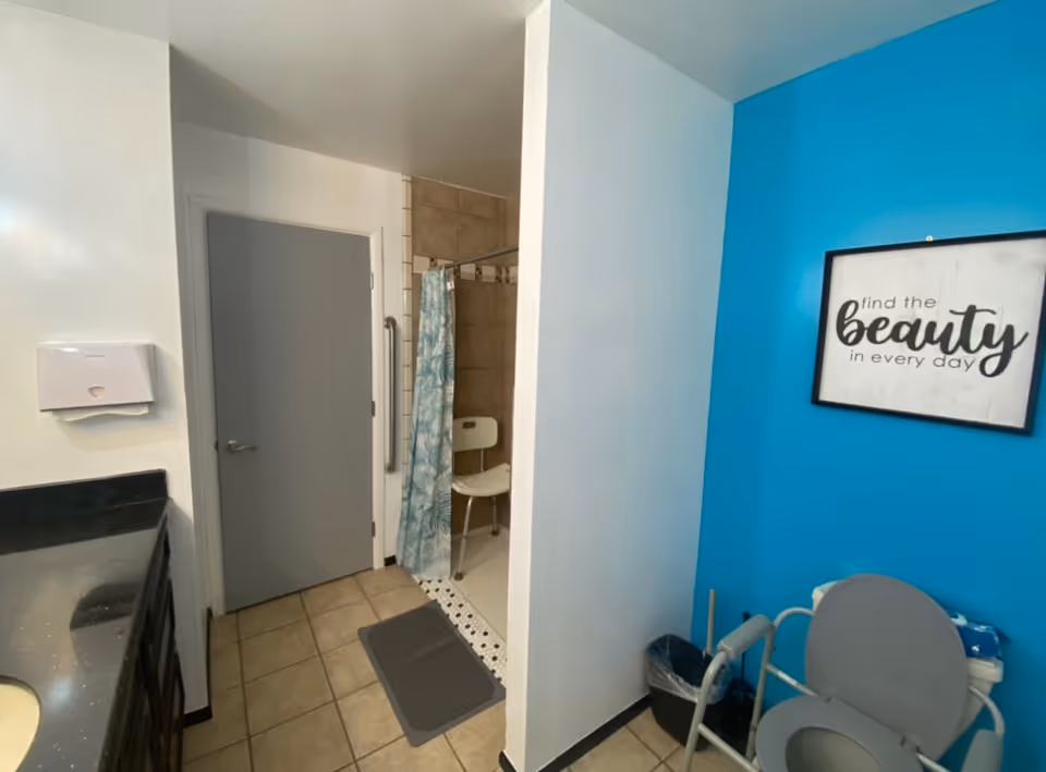 A bathroom with a gray door, a shower area with a blue and white patterned curtain and a white shower chair, a gray toilet with safety rails, a black trash bin, a black countertop with a sink, and a blue wall with a framed sign that reads 'find the beauty in every day'.