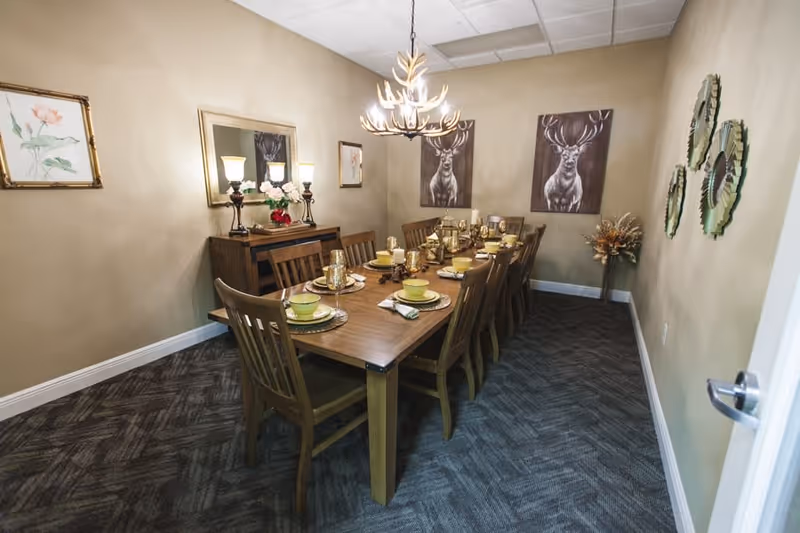 A dining room with a long wooden table set for ten people with matching wooden chairs. The table is set with yellow bowls, plates, and glasses on woven placemats. The room has beige walls decorated with framed artwork, including two paintings of deer and floral prints. A chandelier with antler design hangs above the table, and there is a sideboard with lamps and flowers against one wall. The floor is carpeted with a dark patterned carpet.