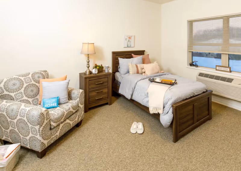 Cozy bedroom with a single wooden bed made with light bedding, a nightstand and lamp, a patterned armchair and slippers on the carpet.