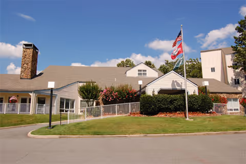 Exterior view of a single-story building with a chimney, surrounded by green grass and bushes. There are three flagpoles with flags in front of the building under a blue sky with some clouds.