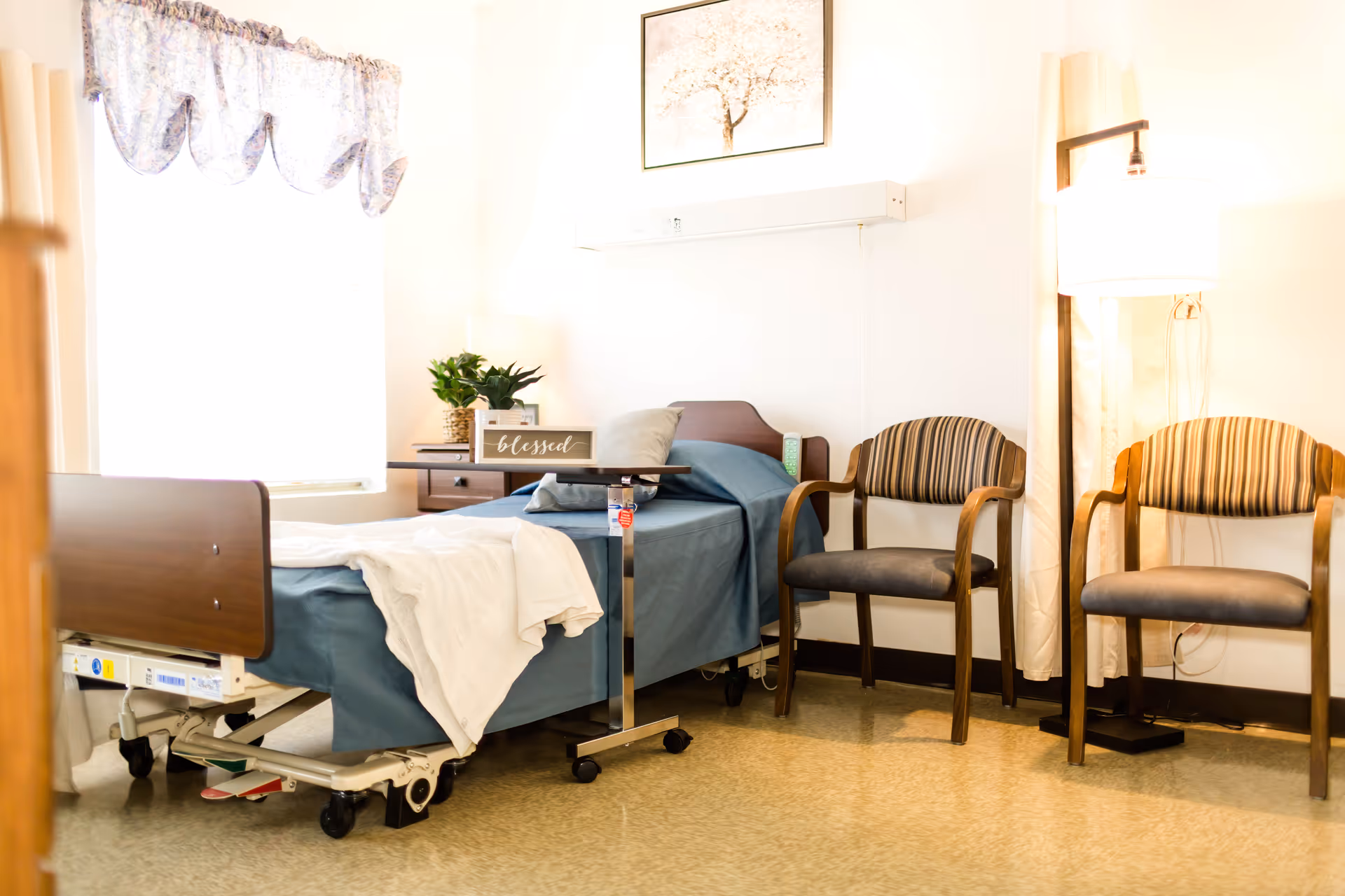 A bright and clean patient room with a hospital bed covered in blue bedding and a white blanket. There is a small overbed table with a decorative sign that says 'blessed' and a plant on a wooden nightstand. Two striped armchairs and a floor lamp are positioned against the wall. A framed picture of a tree hangs above the bed.