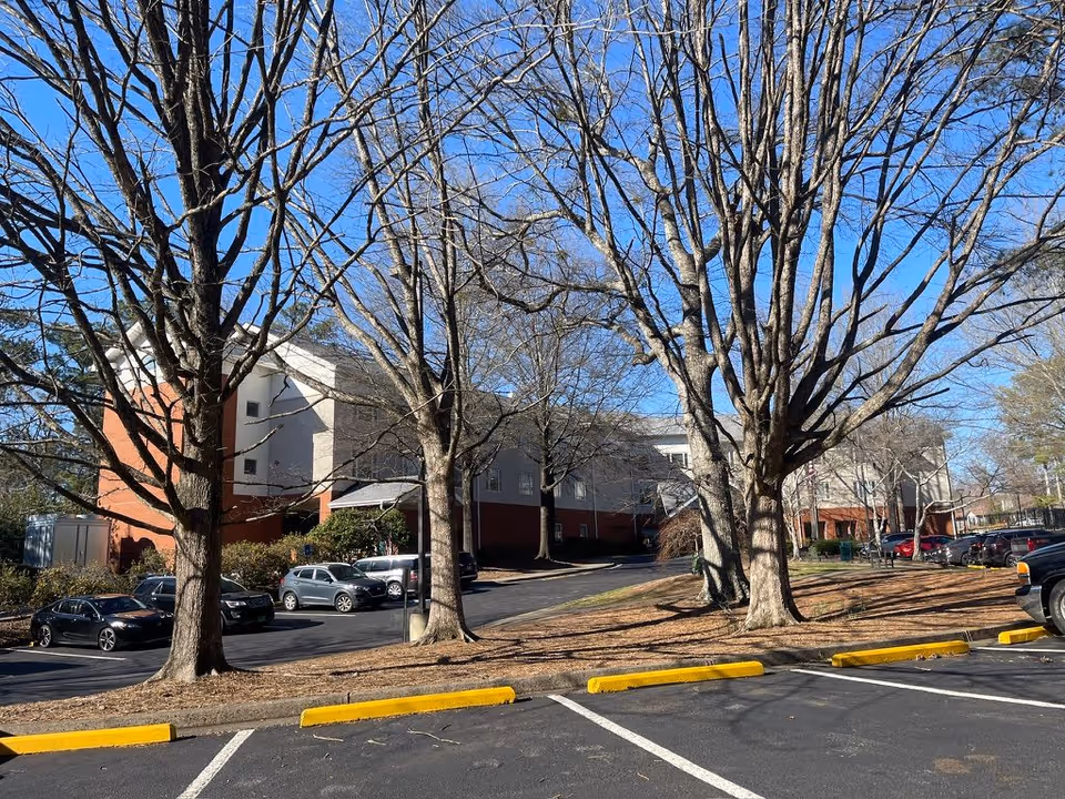 Exterior view of a multi-story brick-and-white senior living building seen behind leafless trees with parked cars in the foreground.