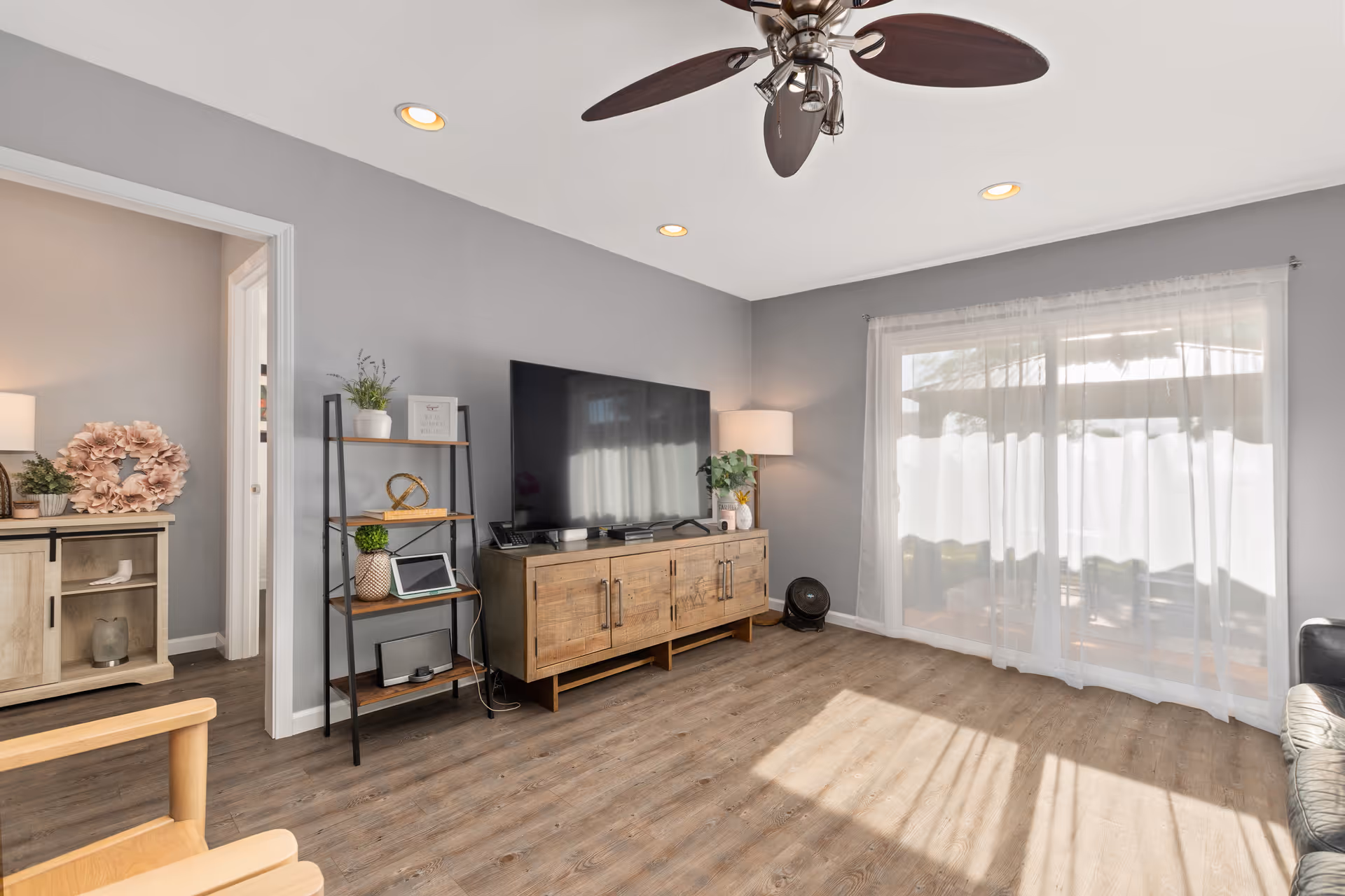 Bright living room with a large TV on a wooden console, a shelving unit, ceiling fan, and sliding glass doors with sheer curtains.