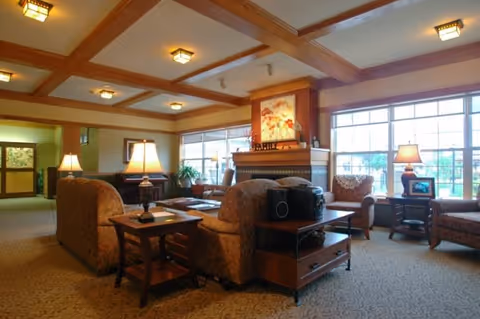 A cozy assisted living community lounge area with patterned upholstered armchairs and sofas arranged around a wooden coffee table and side tables with lamps. The room features a fireplace with artwork above it, large windows letting in natural light, and a ceiling with wooden beams and recessed lighting.
