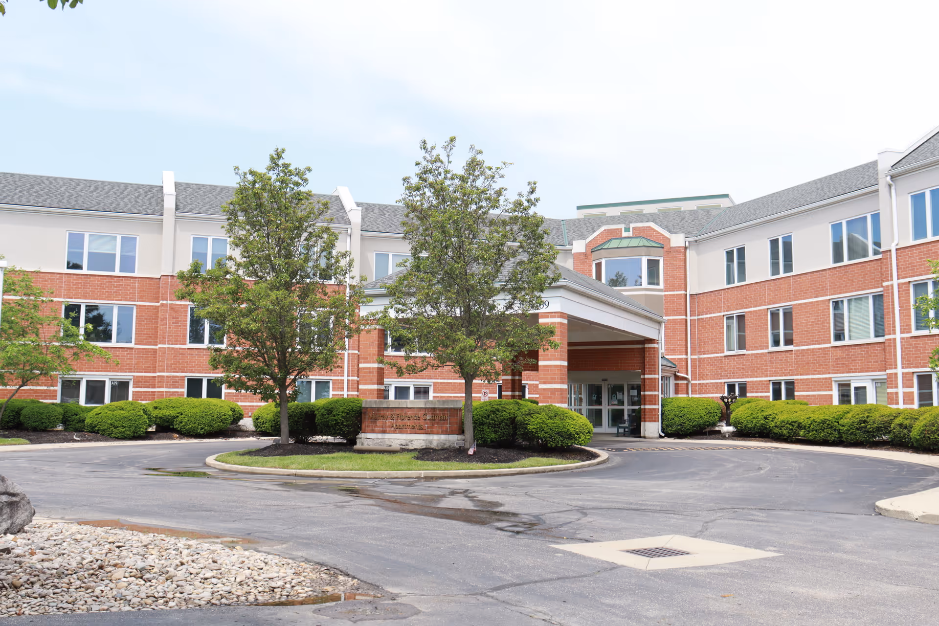 Front exterior view of a three-story senior living facility with red brick and beige siding, featuring a covered entrance, several windows, green bushes, and trees in front.