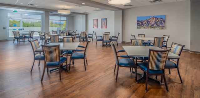 A spacious dining room with multiple square tables, each surrounded by four chairs with striped upholstery. The room has wooden flooring, large windows letting in natural light, and colorful artwork on the walls.