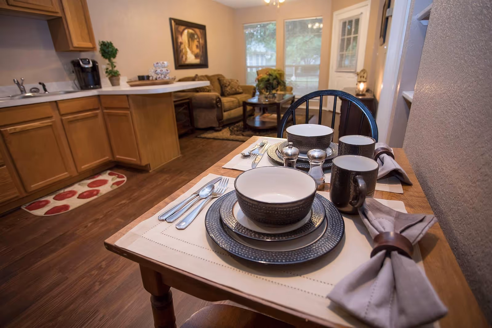 A cozy dining area set for two with dark plates, bowls, mugs, and silverware on beige placemats with cloth napkins in wooden rings. The dining table is adjacent to a kitchen with wooden cabinets and a coffee maker. In the background, there is a living room area with a sofa, coffee table, and a framed painting on the wall.