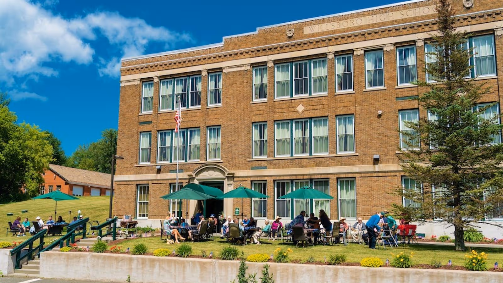 Outdoor scene showing a group of senior individuals sitting and socializing at tables with green umbrellas in front of a large brick building labeled 'Willsboro High School' under a bright blue sky with some clouds.