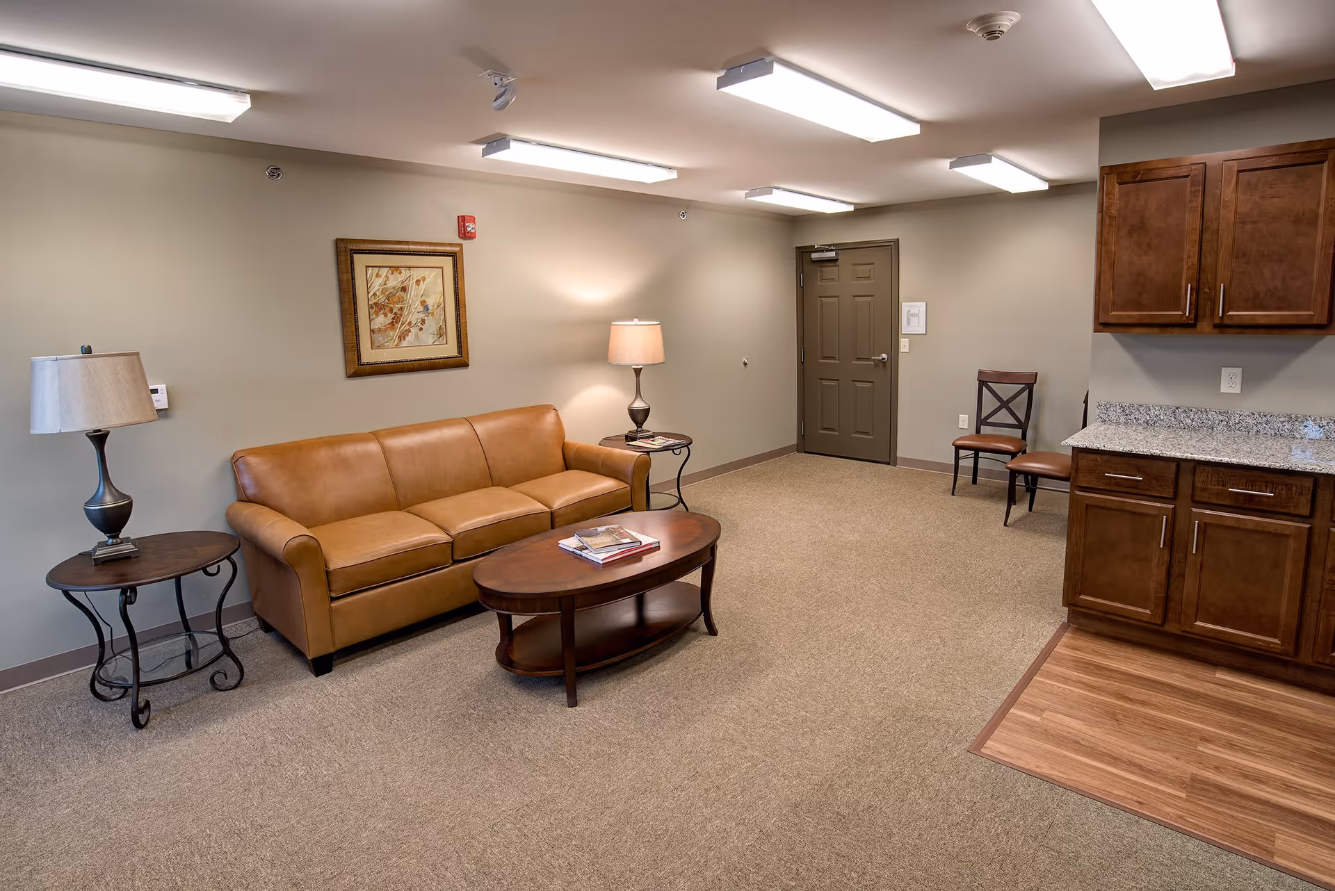 A cozy living room area with a tan leather sofa, two side tables each with a lamp, a wooden coffee table with magazines, a framed artwork on the wall, a closed door, two wooden chairs, and a section of wooden cabinetry with a granite countertop.