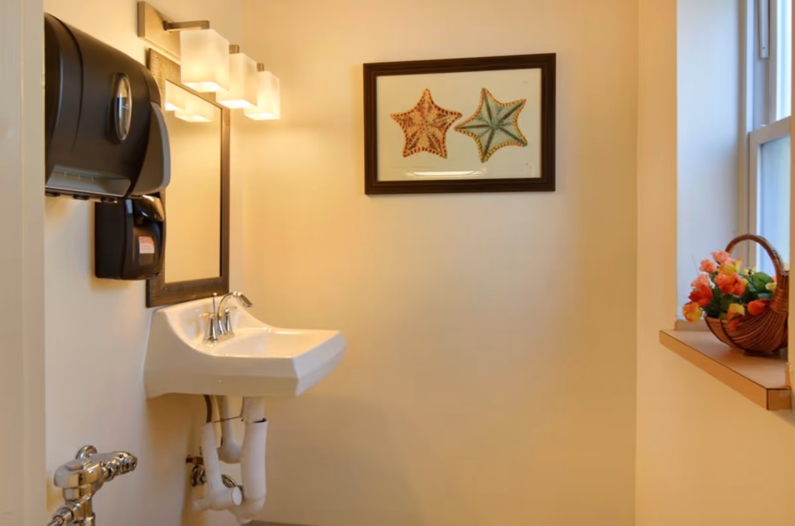 A small bathroom with a white wall-mounted sink, a mirror with three light fixtures above it, a black paper towel dispenser and soap dispenser mounted on the wall, a framed picture of two starfish, and a window sill with a basket of colorful flowers.