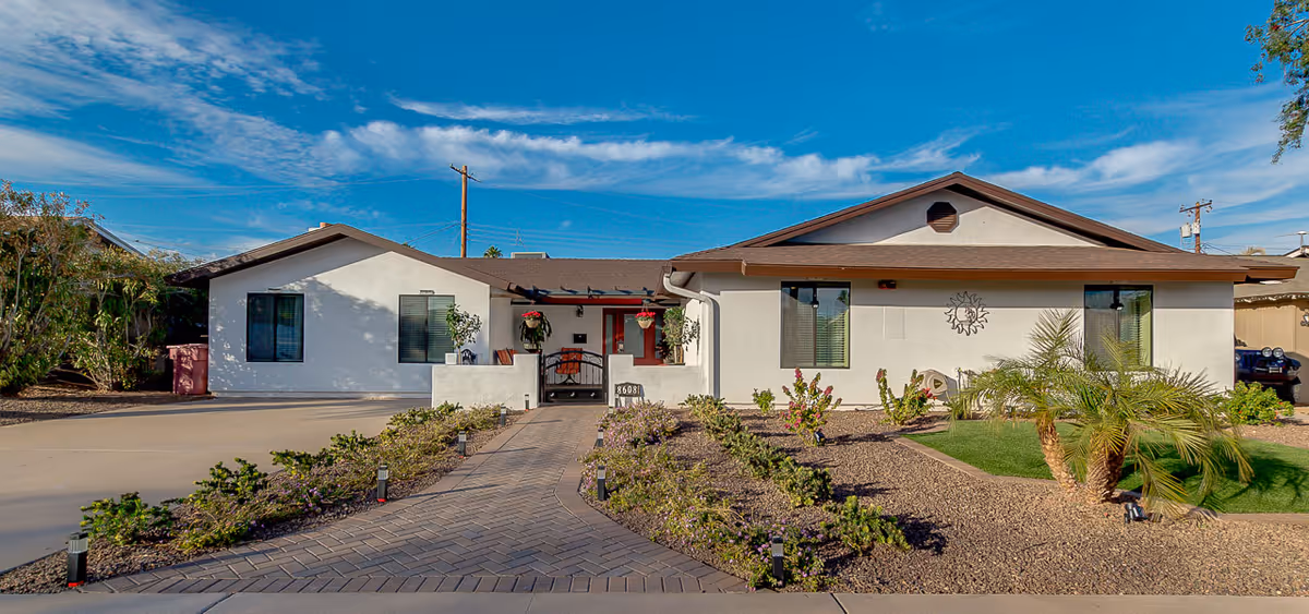 Front exterior view of a single-story assisted living facility building with white walls and brown roof trim. There is a paved walkway leading to the entrance, bordered by small plants and landscaping with gravel and a small palm tree. The sky is blue with some clouds.