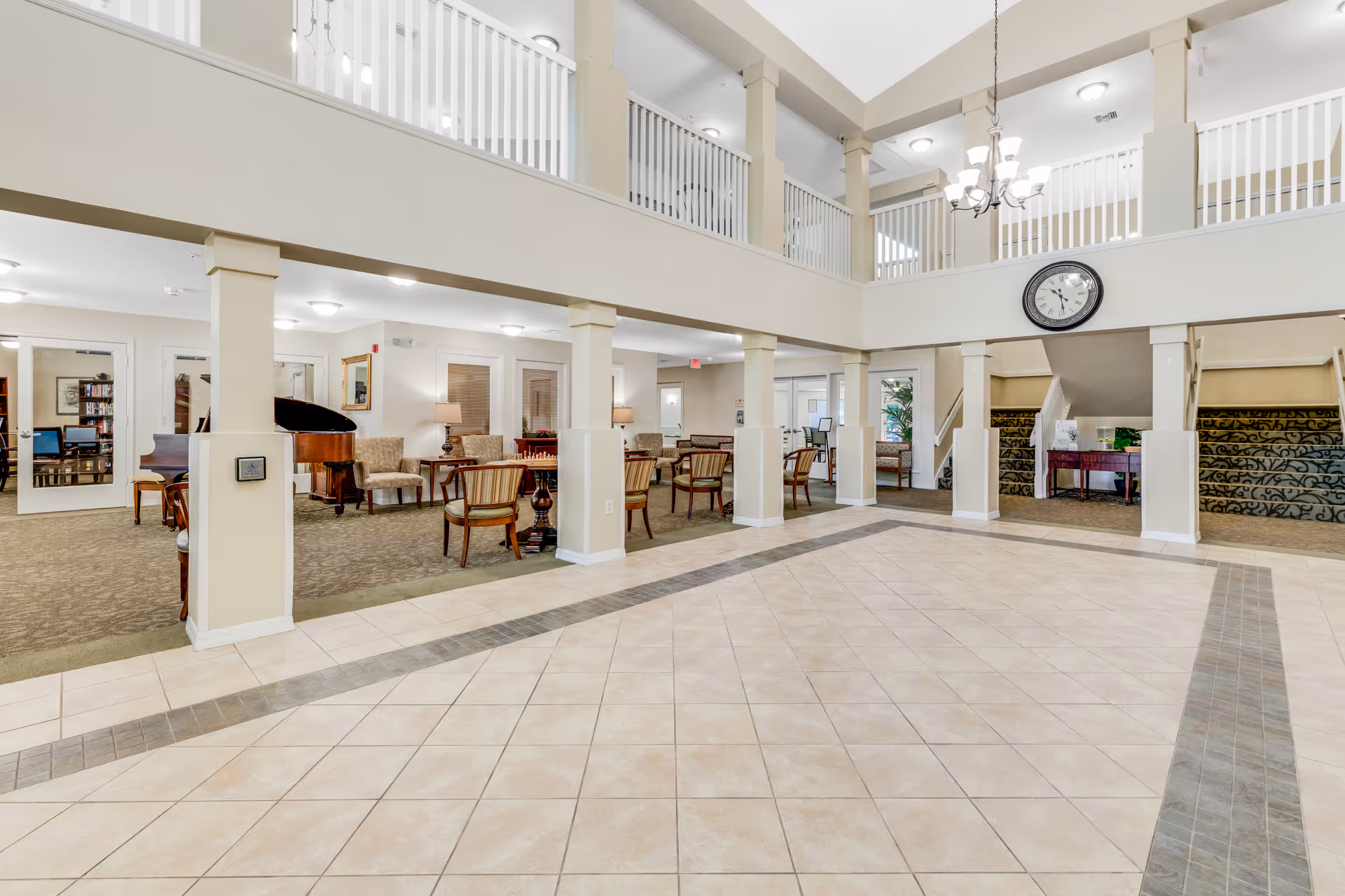 Spacious senior living facility common area with tiled floor and beige walls, featuring a seating area with chairs and tables, a grand piano, a staircase with patterned carpet, and a large clock on the wall above the entrance to the stairs. The area has a high ceiling with a chandelier and a second-floor balcony overlooking the space.