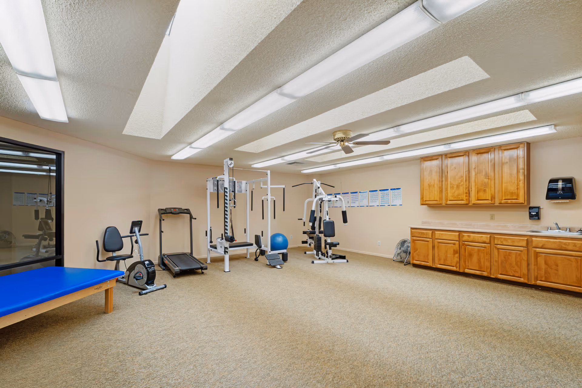 A bright fitness/therapy room with exercise machines, a treadmill, a blue therapy table, and wooden cabinets with a sink under fluorescent ceiling lights.