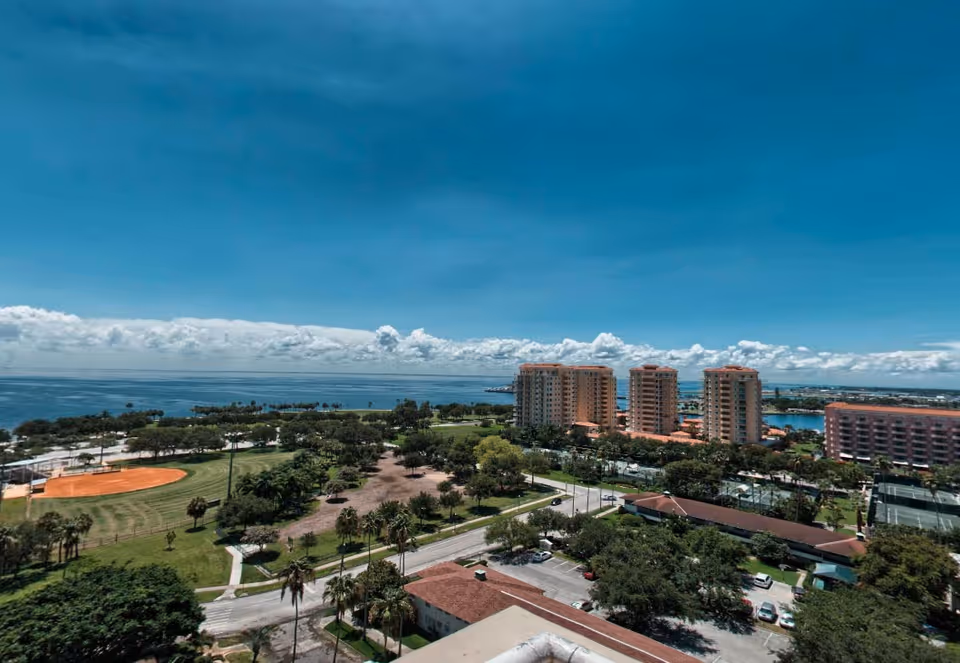 Aerial view of a coastal area featuring a park with a baseball field, several tall residential buildings, a parking lot, and a large body of water under a clear blue sky with scattered clouds.