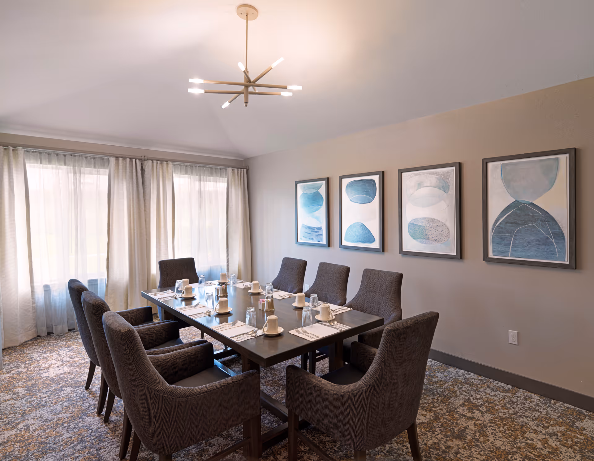 A dining room with a rectangular wooden table set for eight people. The table has white cups, glasses, napkins, and silverware neatly arranged. There are eight upholstered chairs around the table. The room features a modern chandelier hanging from the ceiling and four abstract blue and white paintings on the wall. Large windows with sheer curtains allow natural light to fill the space.