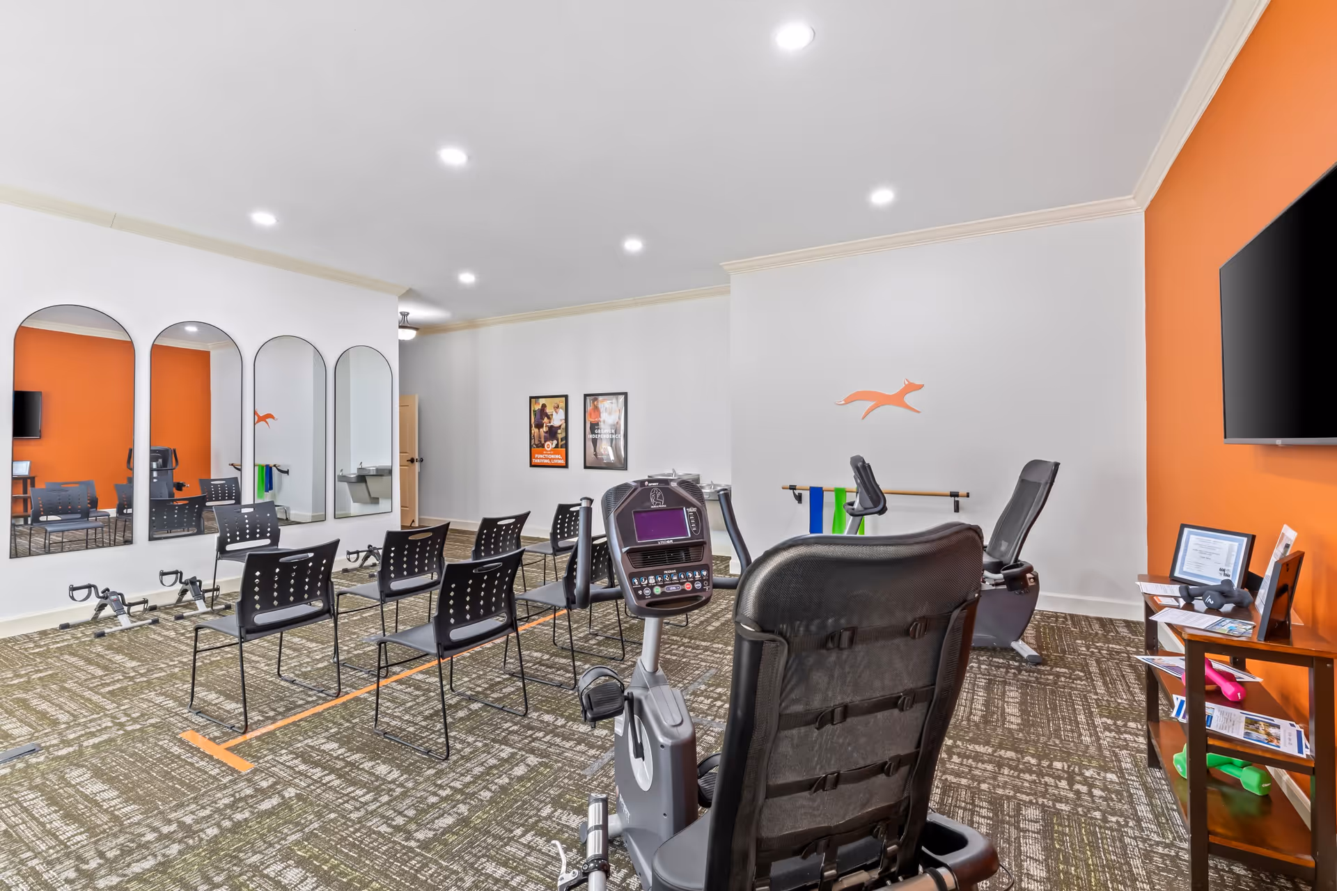 A fitness room in a senior living facility with exercise bikes, chairs arranged in rows, large mirrors on one wall, and an orange accent wall with a mounted TV and a small shelf holding pamphlets and exercise equipment.