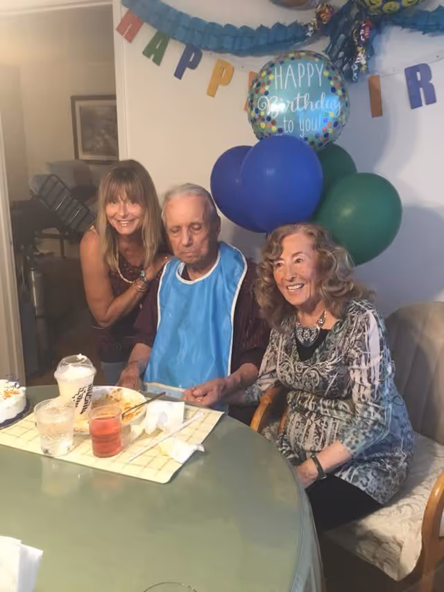 Three people gathered around a table celebrating a birthday. An elderly man wearing a blue bib sits in the center, flanked by two smiling women. Behind them are colorful balloons and a 'Happy Birthday to you!' balloon, along with a partially visible 'Happy Birthday' banner on the wall. On the table are drinks, a plate with food remnants, and a small birthday cake.
