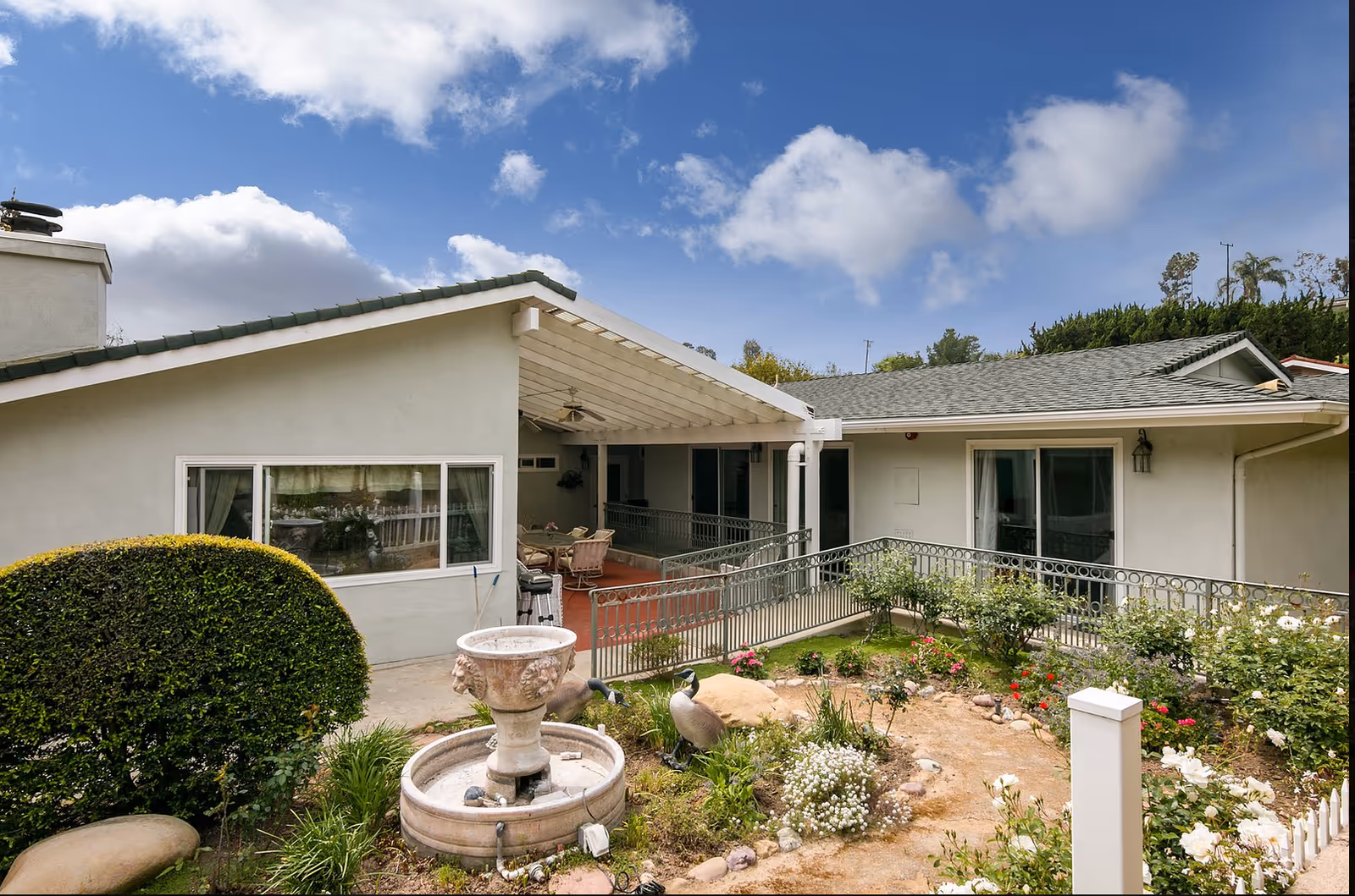 Single-story exterior of a senior living facility with a covered patio, metal railing, garden beds and a decorative fountain under a blue sky.