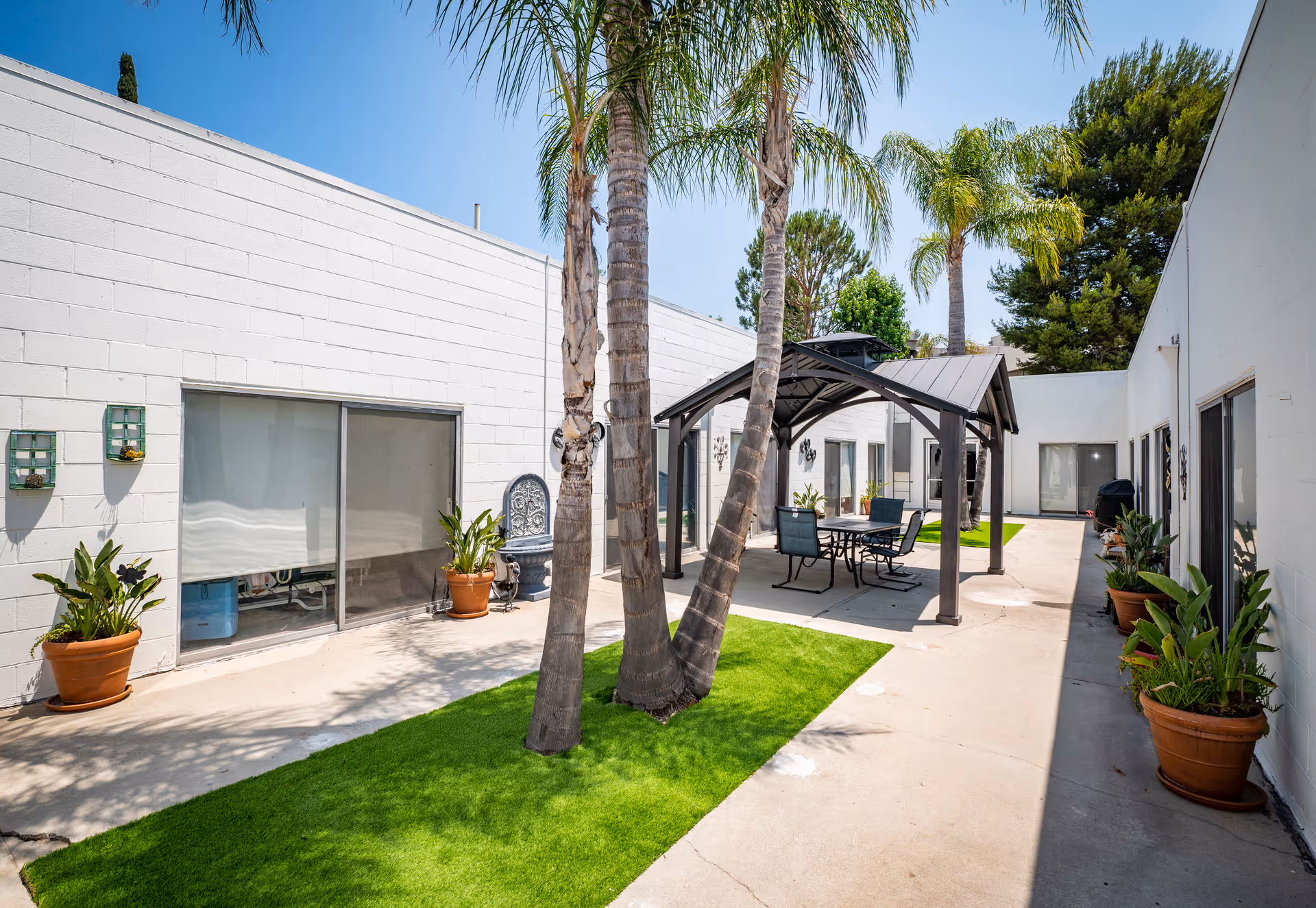Outdoor courtyard area at North Valley Nursing Center with palm trees, potted plants, a covered seating area with a table and chairs, and white building walls with sliding glass doors.